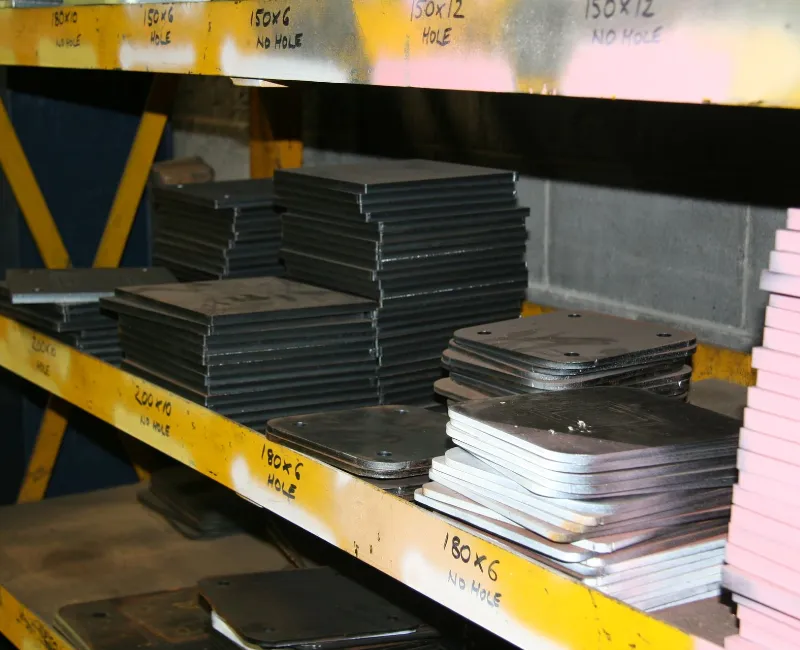 Stacks of steel base plates neatly arranged on yellow industrial shelving in a metal fabrication workshop.