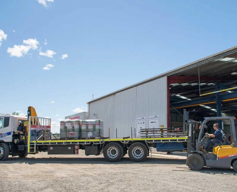 A forklift operator loads steel products onto a flatbed delivery truck outside a large industrial warehouse under a clear blue sky.