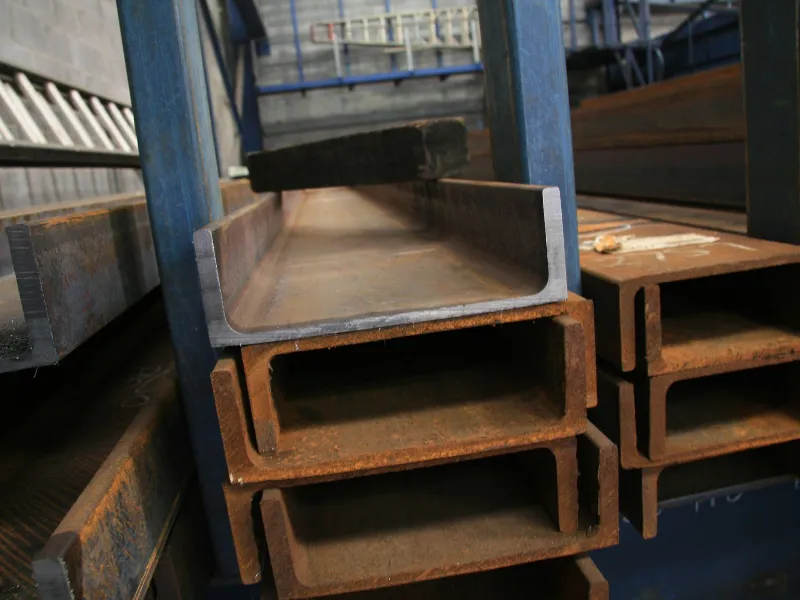 Stacked steel C-sections or channel beams stored in a metal workshop, showing both new and rusted surfaces ready for fabrication or construction use.