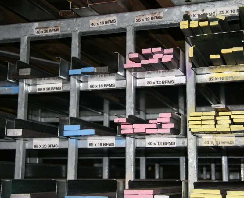 Organised steel flat bars of various sizes and colours stored in labelled metal racks inside a workshop, ready for fabrication or distribution.