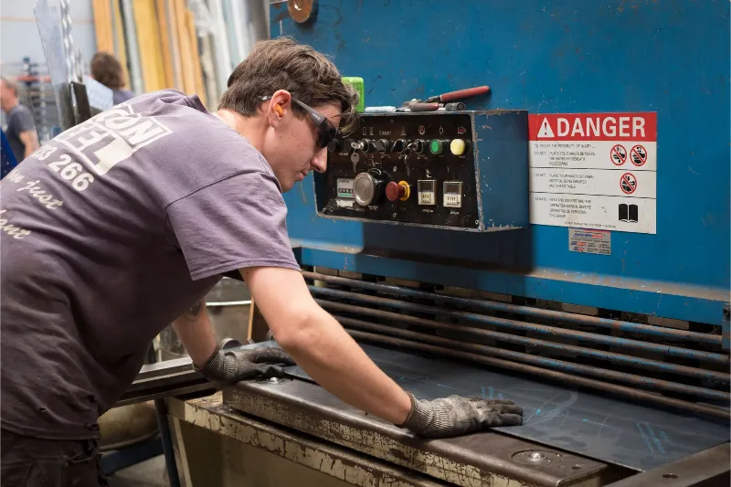 A worker wearing safety glasses and gloves operates a large industrial metal-cutting machine, guiding a steel sheet into place beneath a control panel with warning signs.