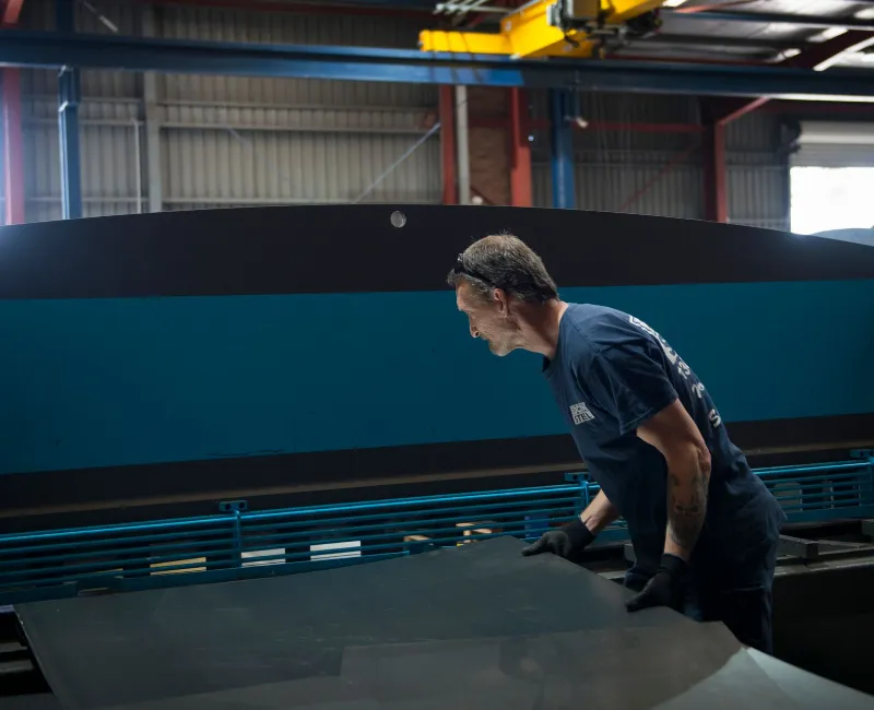 Worker operating machinery to handle a large steel sheet in a fabrication workshop, wearing safety gloves and uniform, with industrial equipment in the background.
