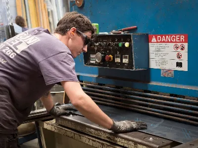 A worker wearing safety glasses and gloves operates a large industrial metal-cutting machine, guiding a steel sheet into place beneath a control panel with warning signs.