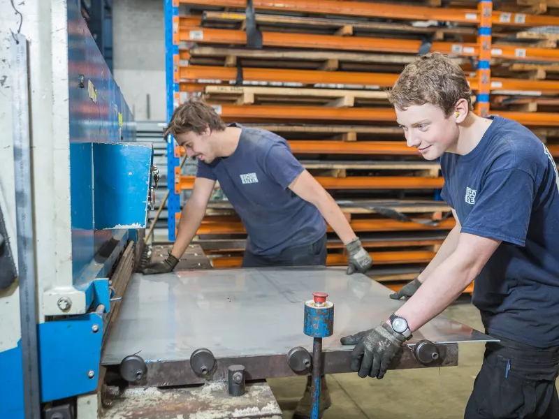 Two workers in a metal fabrication workshop operating a sheet metal cutting machine, wearing safety gloves and navy uniforms, surrounded by metal racks and industrial equipment.