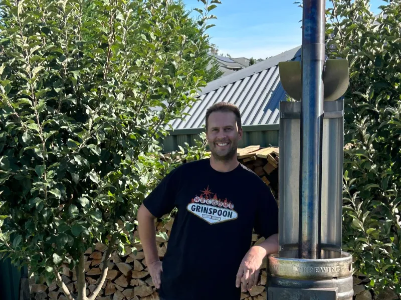 A smiling man standing outdoors beside a tall metal rocket-style outdoor heater, with trees and a shed behind him.
