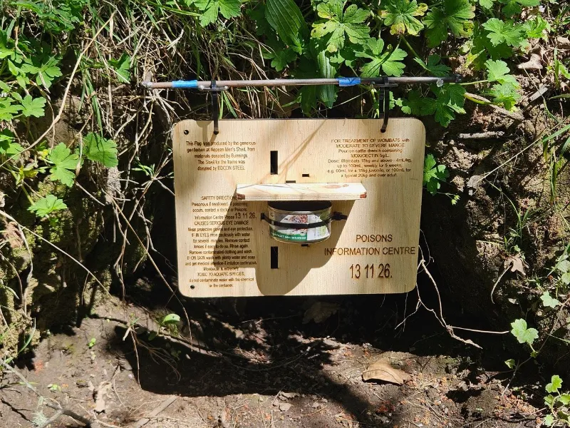 A wooden bait station mounted at ground level in a bushy area, with printed safety information and a container in the centre.