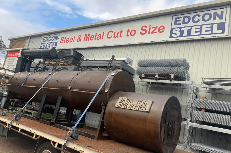 A large “Boss Hog Smokers” steel smoker loaded on a trailer outside an Edcon Steel store, surrounded by metal supplies.