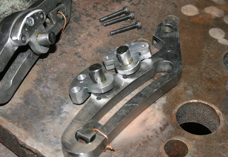 Machined steel brackets and rollers laid out on a workshop bench, with bolts and tools nearby, highlighting raw steel components used in a model locomotive build.