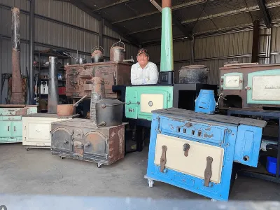 Collection of restored vintage wood stoves in workshop with craftsman standing behind them.
