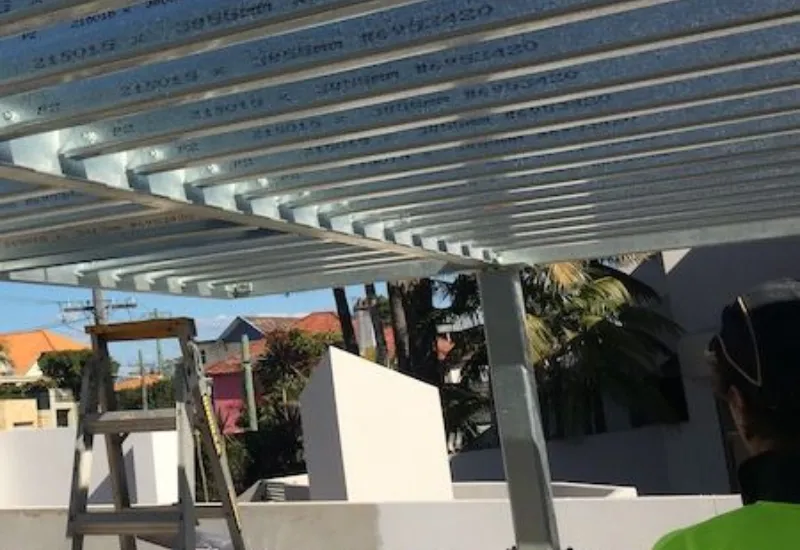 Steel framing being installed on a building under construction; ladder and scaffolding visible in a sunny residential area.