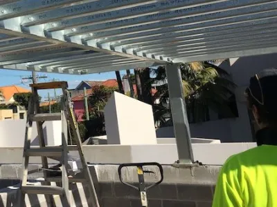 Construction site: overhead metal framing, a ladder propped nearby, and a worker in a neon safety shirt near concrete blocks.