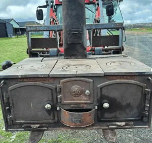 Large vintage wood-fired farm stove mounted outdoors with tractor in background.