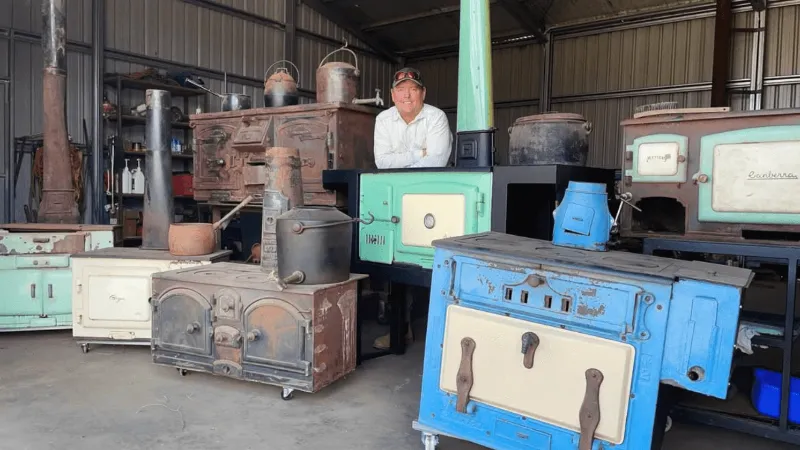 Collection of restored vintage wood stoves in workshop with craftsman standing behind them.