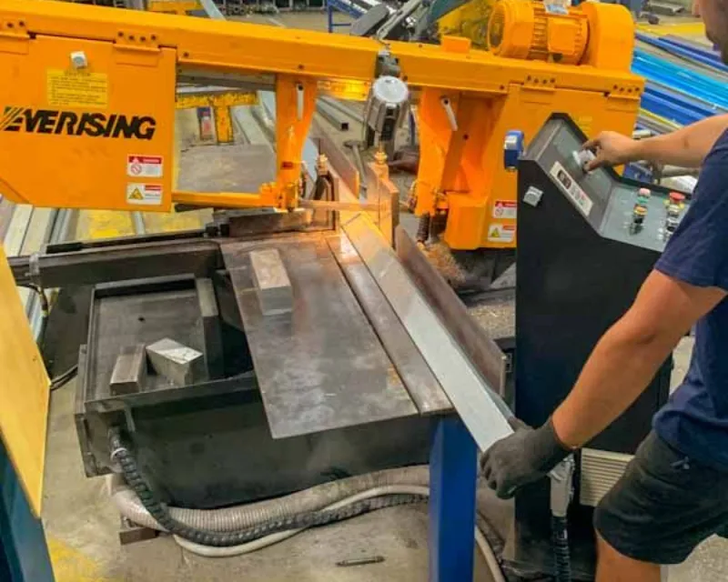 Worker wearing gloves operates an orange metal-cutting saw cutting a steel beam on a fabrication line.