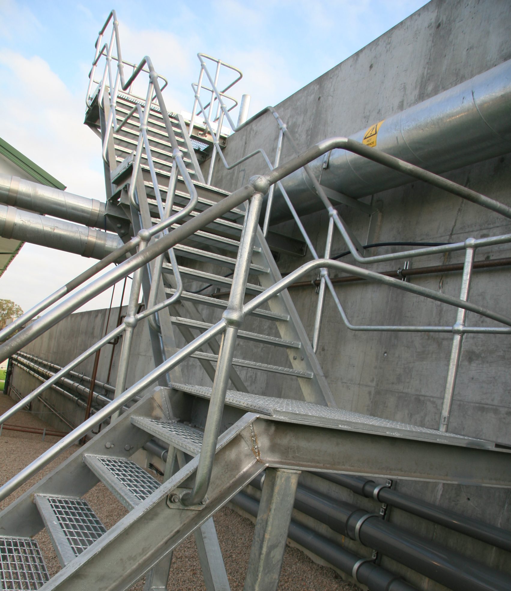 Industrial metal staircase with rails beside large gray pipes along a concrete wall