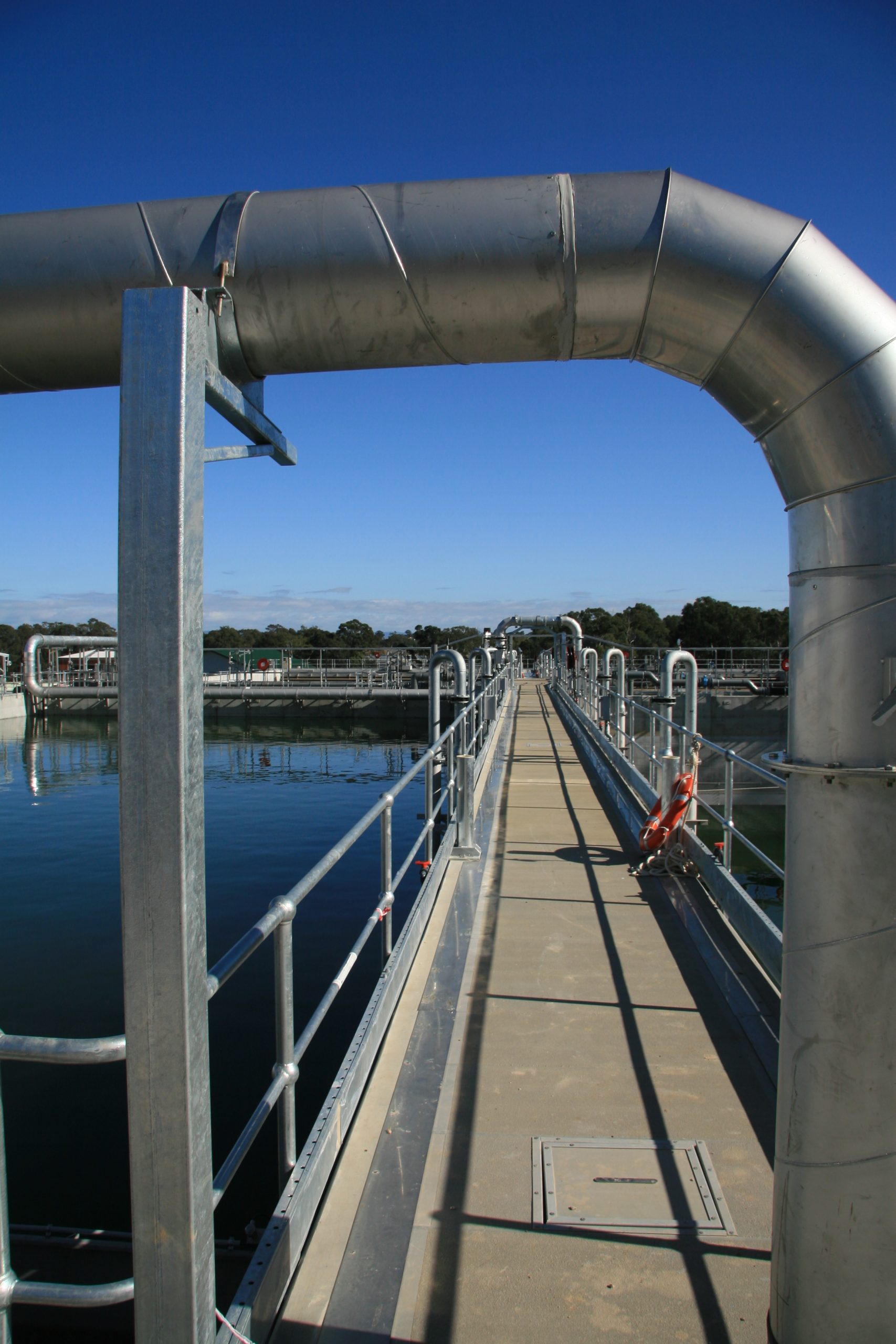 Large curved metal pipe supported by a blue-painted frame runs over a walkway at an industrial dock, with water beneath and clear blue sky above.