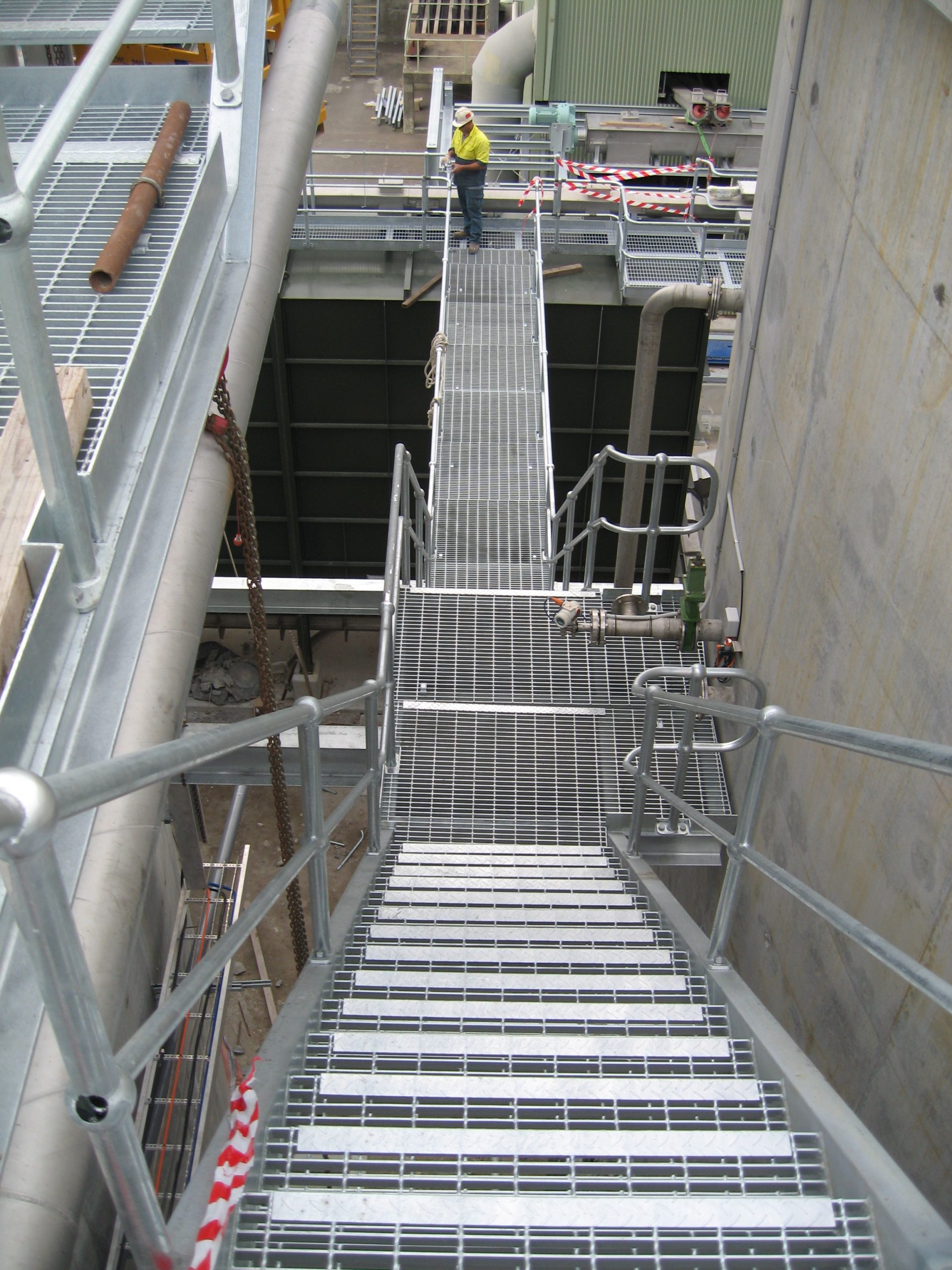Industrial metal stairway over a trench with grated steps and handrails; a worker in a yellow shirt stands on the platform at the top.