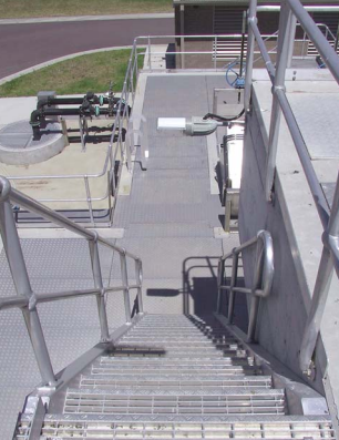 Outdoor metal stairway and catwalk with guardrails at an industrial facility, pipes in the background.