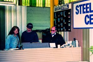 Two customers and a staff member stand at a service counter inside an industrial shop, with a large STEEL sign on the wall behind them.