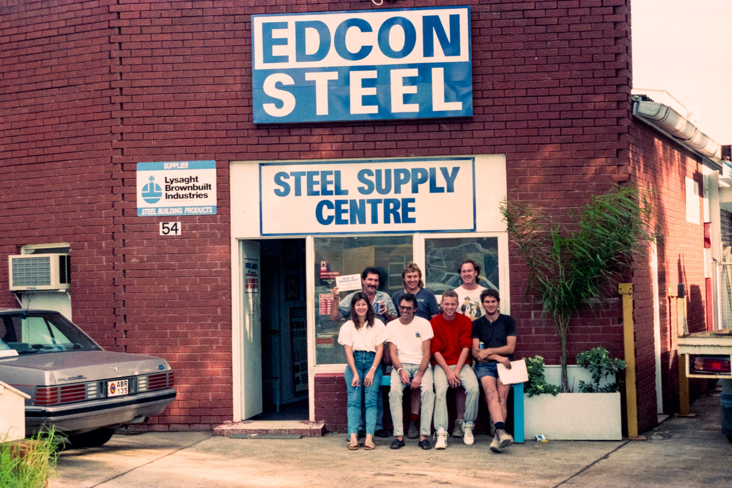 Group of people posing in front of a brick steel supply shop with a large EDCON STEEL sign above and a 'Steel Supply Centre' sign at the entrance.