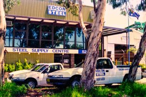 Front of Edcon Steel building with 'STEEL SUPPLY CENTRE' sign, glass façade, and two company vans in the parking lot; trees in foreground.
