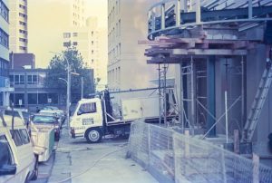 City construction site with scaffolding, wooden planks, and a white Edcon Steel truck in the foreground that reads 'EDCON STEEL' on the door area.