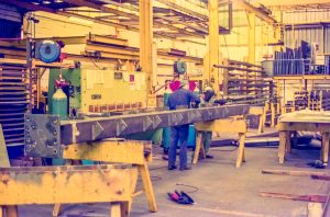 Two workers in blue coveralls weld at a long metal bench in an industrial shop with yellow lighting and equipment nearby