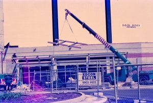 Construction site with scaffolding and a crane lifting metal beams in front of a brick building; 'EDCON STEEL' sign visible on a banner.