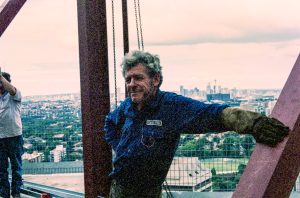 Smiling man in a blue shirt stands on a high metal platform with chains, overlooking a city skyline in the distance.