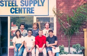 Group of people posing in front of a brick storefront with a 'STEEL SUPPLY CENTRE' sign above them and greenery nearby.