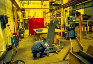 Worker in protective gear welding a metal piece at a cluttered industrial workshop.