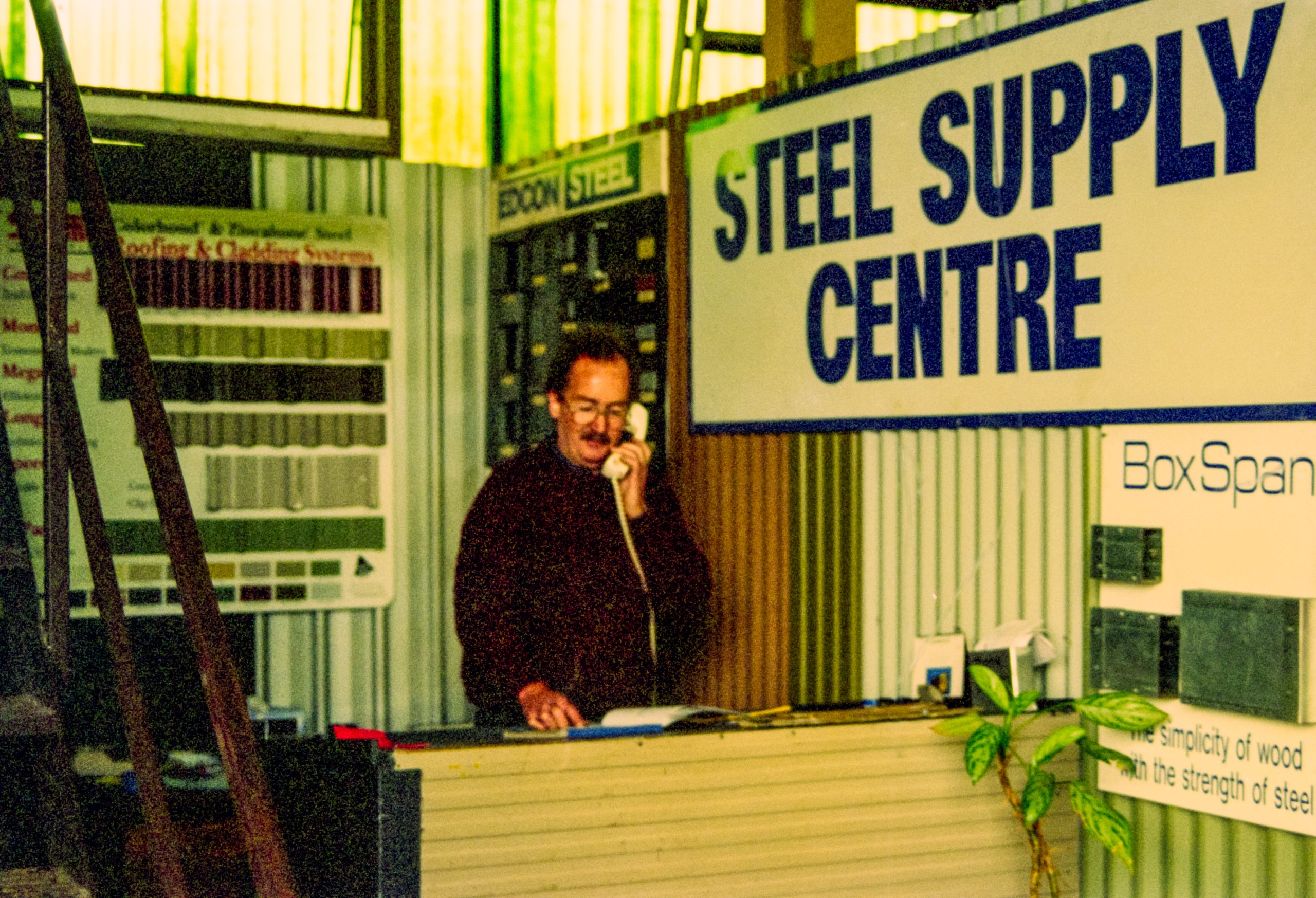 Man in a dark sweater on a landline phone at a steel supply shop counter, with a large 'STEEL SUPPLY CENTRE' sign in the background.