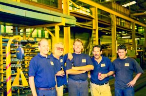Group of five factory workers in blue uniforms posing together in a yellow steel-structured warehouse.