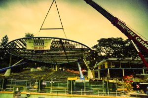 Under-construction roller coaster with blue track and a crane lifting a support beam over empty stadium-style seats