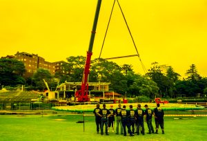 Line of construction workers in dark shirts stand in a row watching a large red crane lift a beam at an outdoor construction site with buildings behind them.