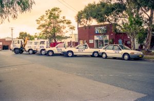 Row of white Edcon Steel fleet trucks parked along a street in front of a brick building with trees overhead (industrial scene)