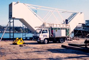 Construction worker stands on top of a large white modular structure mounted on a truck, with metal scaffolding and a crane-like frame on a rooftop.