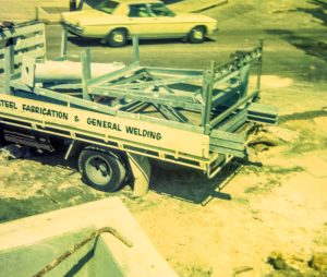 Industrial metal trailer on a dusty site, loaded with scaffolding and metal frames, with a sign reading 'Steel Fabrication & General Welding' along the side.