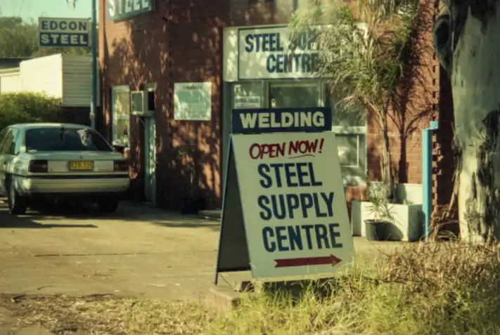 Signboard outside a steel supply centre advertising 'Open Now! Steel Supply Centre' with a brick building, palm trees, and a parked car nearby.