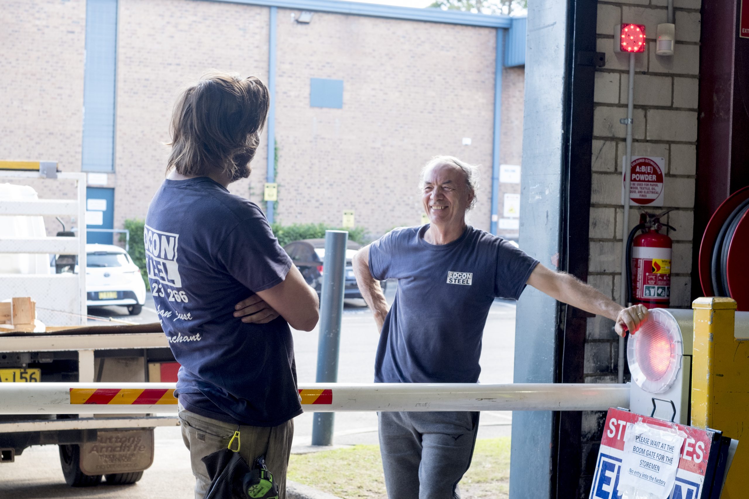 Two men in blue shirts chat beside a barrier at a loading dock area, smiling as they talk.