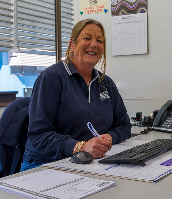 Smiling woman in a navy polo shirt sits at a desk, writing with a pen beside a computer keyboard and mouse while a calendar hangs on the wall behind her.