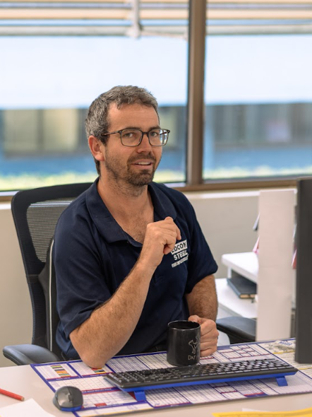 Man with glasses sitting at office desk, holding a mug and looking at the camera, wearing a navy polo with a company logo, with a keyboard and monitor nearby.